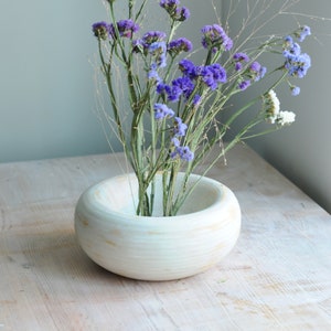 May include: A white ceramic bowl with a rounded shape and a textured surface holds a bouquet of purple and white flowers. The flowers are arranged in a simple, elegant style.