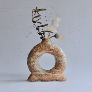 May include: A ceramic vase with a ring-shaped opening and a speckled brown and white glaze. The vase has three legs and is holding dried flowers.