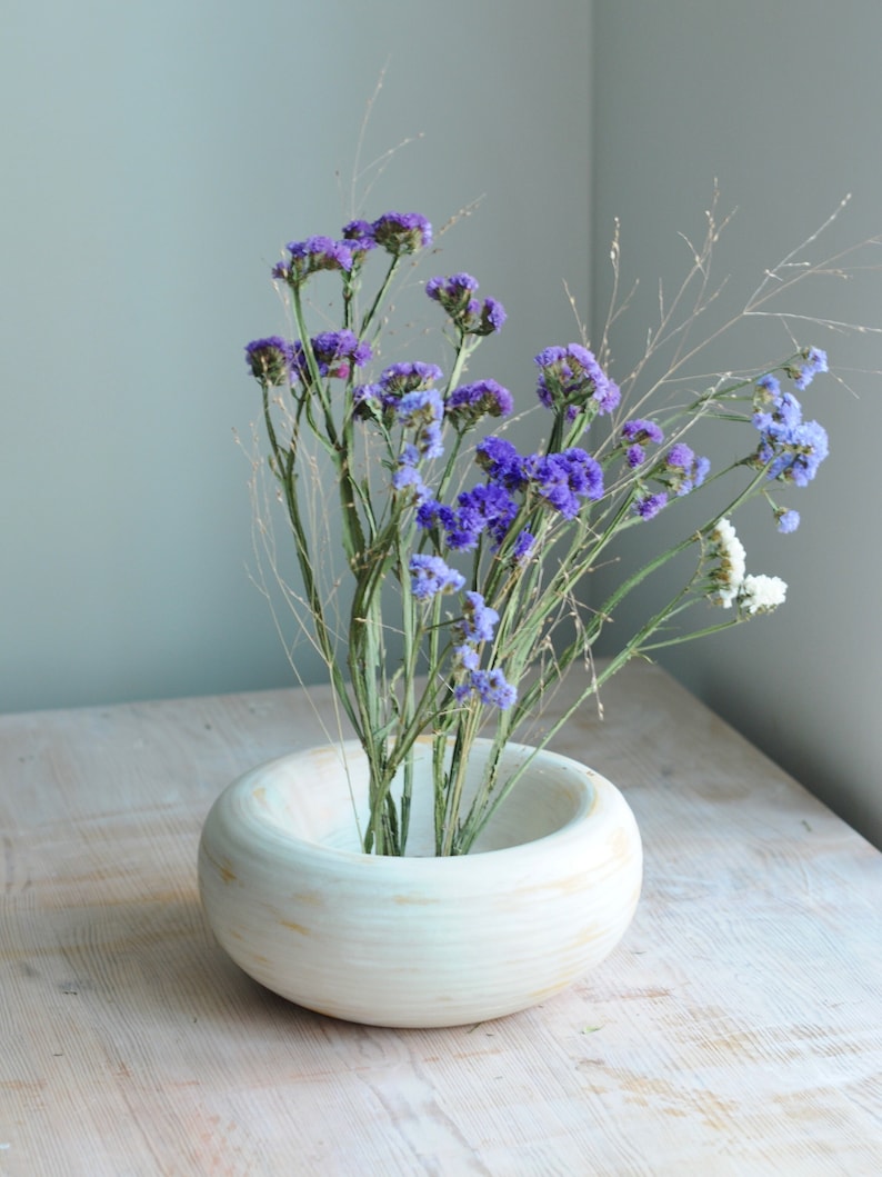 May include: A white ceramic bowl with a rounded shape and a textured surface holds a bouquet of purple and white flowers. The flowers are arranged in a simple, elegant style.