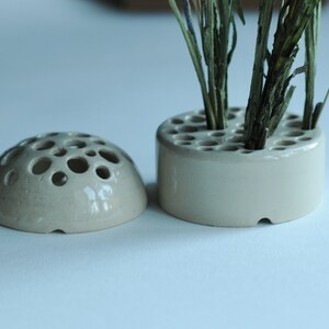May include: Two ceramic flower frog vases, one round and one dome-shaped, with multiple holes for holding stems. The round vase has dried plant stems in it.