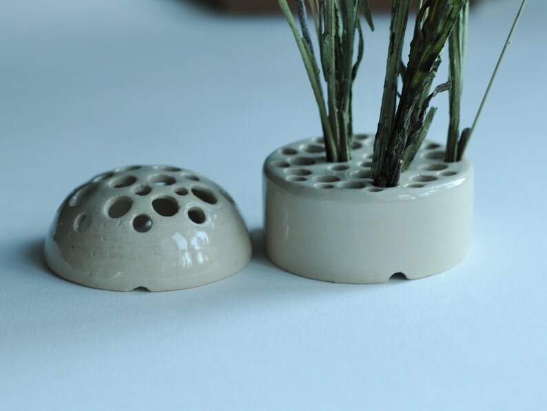 May include: Two ceramic flower frog vases, one round and one dome-shaped, with multiple holes for holding stems. The round vase has dried plant stems in it.