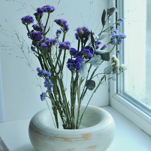 May include: A white ceramic vase with a wide, round opening, featuring a rustic, wood-grain design. The vase is filled with a bouquet of dried purple flowers and green foliage.