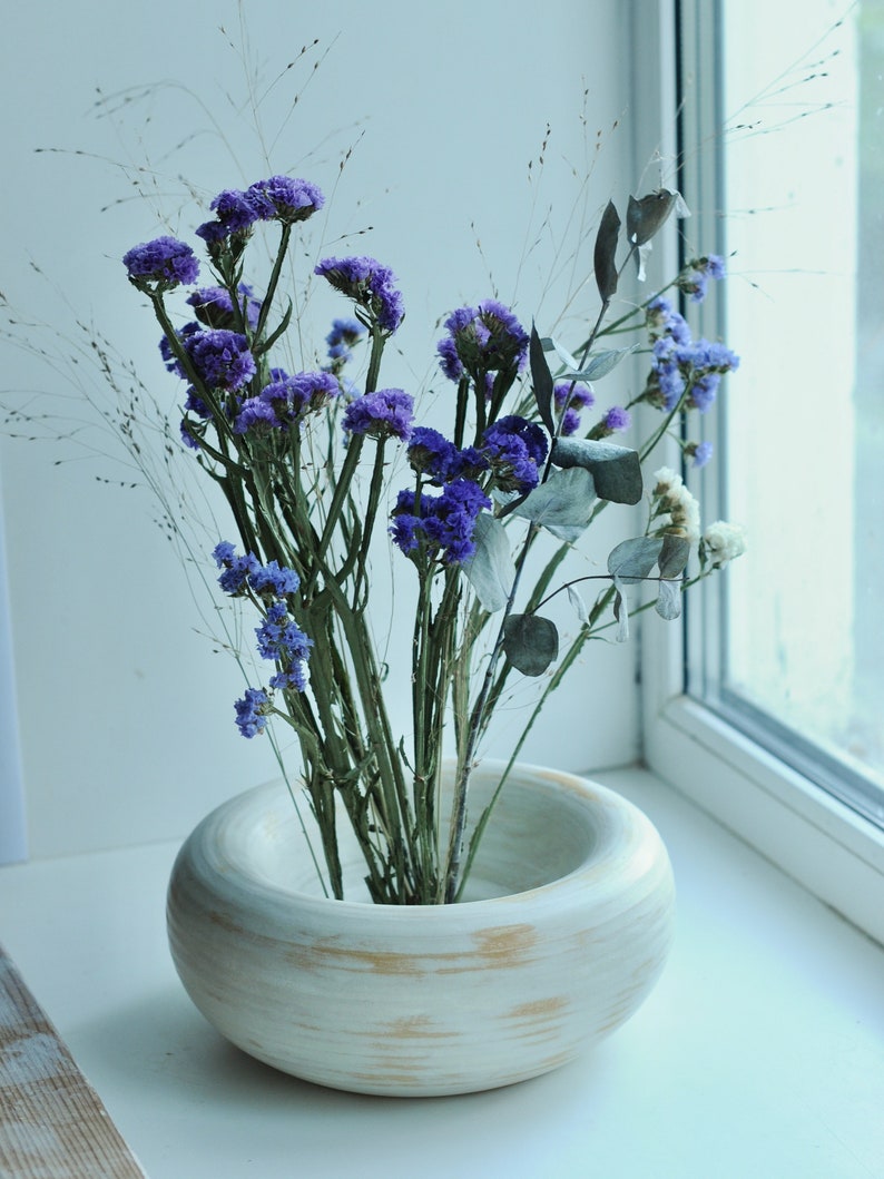 May include: A white ceramic vase with a wide, round opening, featuring a rustic, wood-grain design. The vase is filled with a bouquet of dried purple flowers and green foliage.