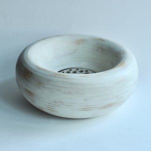 May include: A white ceramic bowl with a round shape and a small hole in the center. The bowl has a distressed white finish with subtle brown accents.