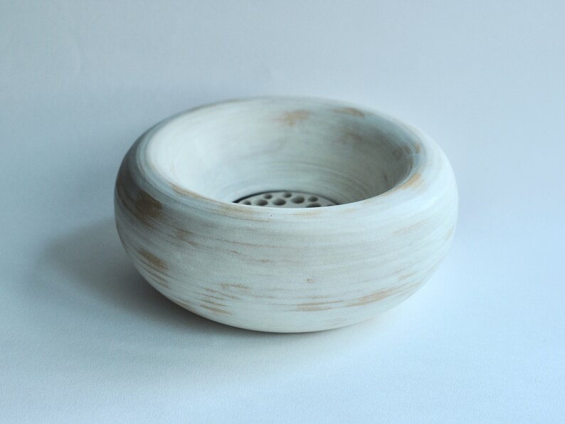 May include: A white ceramic bowl with a round shape and a small hole in the center. The bowl has a distressed white finish with subtle brown accents.