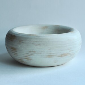 May include: A white ceramic bowl with a distressed finish. The bowl has a rounded shape and a smooth, white glaze. The glaze has a subtle, distressed look, with hints of brown showing through.