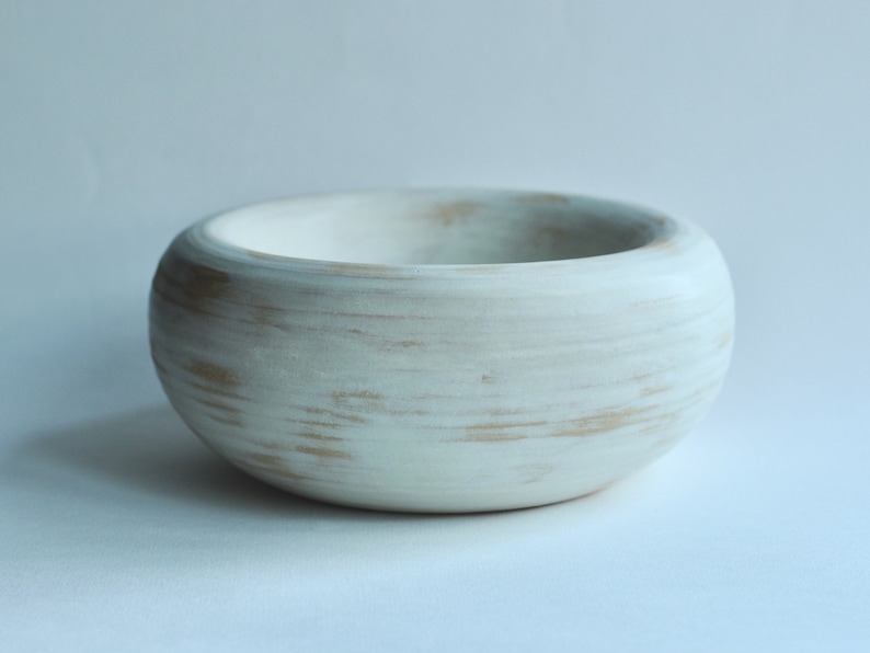 May include: A white ceramic bowl with a distressed finish. The bowl has a rounded shape and a smooth, white glaze. The glaze has a subtle, distressed look, with hints of brown showing through.