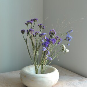 May include: A white ceramic vase with a wide, round opening, filled with a bouquet of purple and white flowers. The vase is sitting on a light wood table.