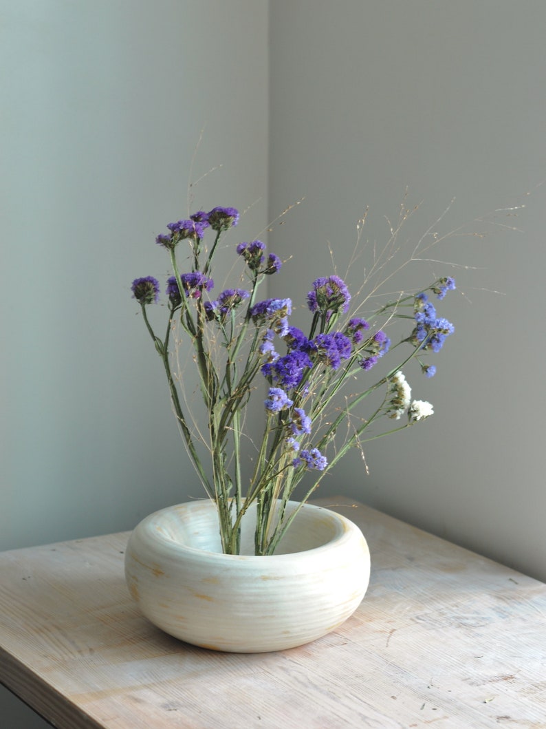May include: A white ceramic vase with a wide, round opening, filled with a bouquet of purple and white flowers. The vase is sitting on a light wood table.