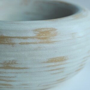 May include: A white ceramic bowl with a light brown glaze. The glaze has a streaky, textured appearance.