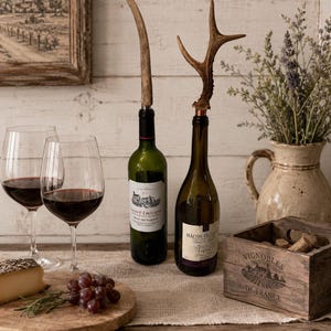 May include: Two wine bottles topped with natural deer antler stoppers. One bottle features a Saint-Emilion label, while the other displays a Macon-Villages label. The stoppers are crafted from authentic brown antler tines, providing a rustic accent to the glass bottles. These decorative bottle toppers are designed to fit standard wine bottle openings, showcasing a distinct organic texture and shape on each piece.