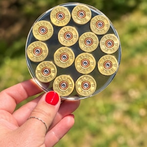 May include: A circular clear coaster embedded with thirteen brass-coloured ELEY 12 gauge shotgun shell heads. Each metallic base features the text ELEY and the number 12 stamped into the surface, arranged in a pattern within the transparent resin disc.