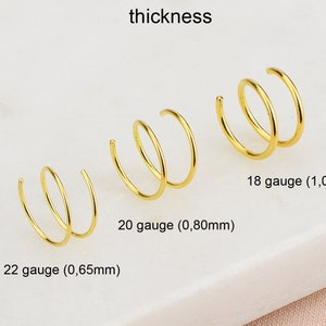 May include: Three gold nose rings of different thicknesses, labelled with their gauge and diameter in millimetres. The rings are arranged from largest to smallest, with the largest ring on the right and the smallest ring on the left.