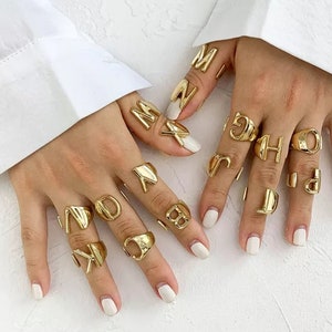 Puede incluir: Un conjunto de anillos de color dorado con diferentes letras del alfabeto. Los anillos se llevan en varios dedos de una mano.