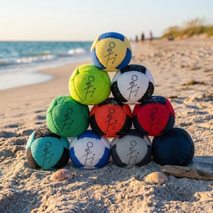 May include: A pyramid of colorful, small fabric balls on a sandy beach. The balls are in various colors, including yellow, green, red, and blue, each with a stylized figure printed on them. The ocean and sky are visible in the background.