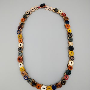 May include: A handmade necklace featuring a variety of colorful buttons in shades of red, blue, yellow, and brown. The buttons are attached to a light-colored, woven base, creating a unique and artistic accessory. The necklace is displayed against a plain gray background.