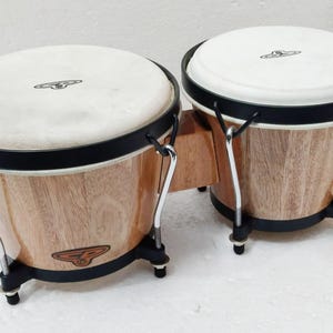 Puede incluir: Un par de tambores bongo conectados con cascos de madera de color marrón claro con un acabado de veta natural. Los tambores tienen parches blancos, aros de metal negro y tensores plateados. Un pequeño logo con las letras CP es visible.