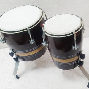 May include: A pair of bongo drums with dark brown shells, white drumheads, and metallic hardware. Each drum features a horizontal gold stripe accented by thin white lines. The drums are mounted on individual metal stands with black rubber feet.