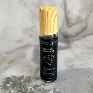May include: A 10 ml roll-on bottle of essential oil blend with a black label that says "Labradorite Infused Essential Oil Blend", "Foxwand Apothecary", and "Lightworker for Spiritual Growth & Transformation". The bottle has a wooden cap and is sitting on a bed of small, clear stones.