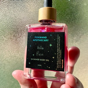 May include: A 50 ml bottle of shimmer body oil labelled "Foxwand Apothecary Hello Pixie Shimmer Body Oil". The bottle is clear glass with a pink liquid inside and a wooden cap with a black dropper.