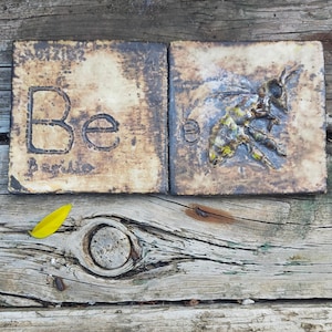 Puede incluir: Dos azulejos de cerámica con un esmalte rústico marrón y beige. El azulejo de la izquierda tiene la letra "Be" en letras negras. El azulejo de la derecha tiene una imagen estilizada de una abeja en amarillo, marrón y blanco.