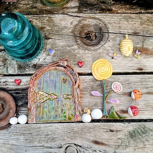 May include: A ceramic fairy door with a green door and a brown, tree-like frame, featuring a heart-shaped detail. Decorative ceramic pieces include a yellow flower, ladybirds, and a beehive. The scene is set on a weathered wooden surface.