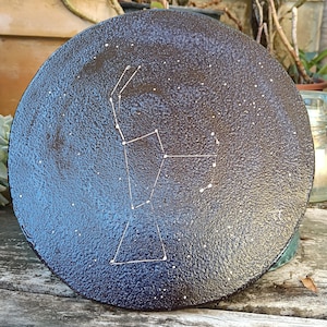 Op de afbeelding: Een ronde, donkerblauwe decoratieve plaat met een textuur. De plaat heeft een wit sterrenbeeldontwerp met kleine witte stippen die sterren voorstellen. De plaat heeft een diameter van ongeveer 25 cm.