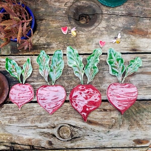 Puede incluir: Cuatro adornos de cerámica con forma de remolacha con hojas verdes. Las remolachas son de color rojo intenso y tienen un acabado brillante. Los adornos están dispuestos en fila sobre una superficie de madera.