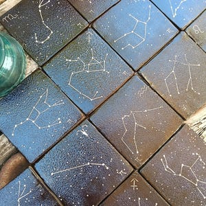 May include: A collection of square tiles with a dark, speckled background, each featuring a white constellation design and symbol. The tiles are arranged together, creating a celestial theme. A glass jar is visible in the upper left corner.