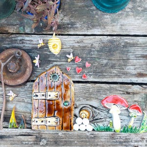 May include: A ceramic fairy door with a brown wooden door, a green window, and two metal hinges. The door is surrounded by red hearts, yellow bees, and white mushrooms.
