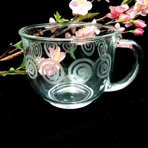 May include: A clear glass teacup with a handle, decorated with etched spiral designs. Pink and white flowers and green leaves are arranged around the cup. The background is black.