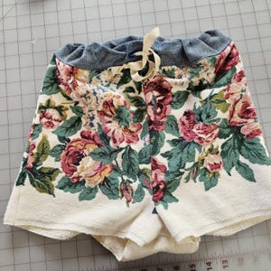 May include: Floral print shorts with a cream-colored base and a pattern of red, pink, and green roses. The waistband is a faded blue with a drawstring closure. The shorts are made of a soft, absorbent material.