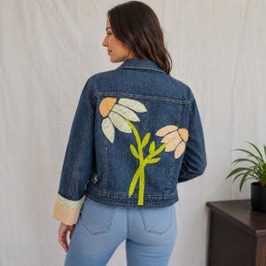May include: A denim jacket with large, embroidered daisy flowers on the back. The flowers have white, peach, and yellow accents, with a green stem. The cuffs are a light peach color with a lace trim. The jacket is a medium wash blue.