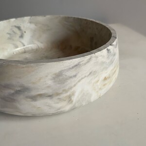 May include: A round, marbled bowl with a smooth interior and a thick, textured rim. The bowl features a swirling pattern of white, grey, and tan hues, creating a natural, stone-like appearance. This decorative bowl is suitable for various uses.