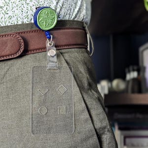 May include: A clear plastic badge holder with a blue and green retractable badge reel attached to a brown belt. The badge holder has four empty slots for inserting different shapes.