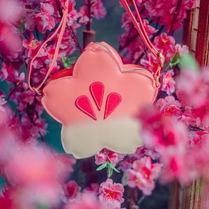 Puede incluir: Un bolso de hombro de color rosa y blanco con forma de flor y una correa rosa. El bolso tiene un diseño de flor rosa en la parte delantera.