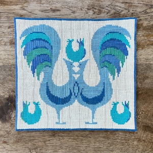 May include: A square needlepoint canvas featuring two large roosters facing each other in shades of blue and teal. The design includes a smaller rooster centered above and two small rooster motifs at the bottom corners, all on a cream background.