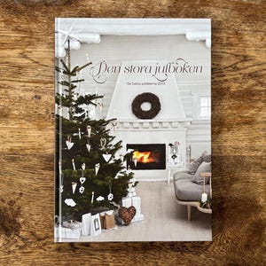 Peut inclure: Un livre relié intitulé Den stora julboken avec une couverture illustrée d'un sapin de Noël décoré, une cheminée avec un feu allumé, une couronne sombre sur la cheminée et un canapé gris dans une pièce lumineuse. Le texte est en blanc.