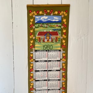 May include: A vintage 1980 calendar printed on fabric with a green background and a floral border. The calendar features a colorful illustration of a house with a red roof and a blue sky. The months of the year are listed in Swedish.