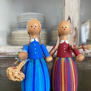 May include: Two wooden dolls with painted faces and hair. The doll on the left is wearing a blue and white striped dress and holding a woven basket. The doll on the right is wearing a red and yellow striped dress.