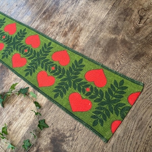 May include: A green runner with a repeating pattern of red hearts and dark green leaves. The runner is rectangular and appears to be made of a woven material. The runner is approximately 60 inches long.
