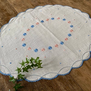 May include: Oval white linen doily with a scalloped blue crochet edge. The doily features an embroidered floral design in a ring of blue and pink flowers. Small blue and pink dots are scattered across the surface.