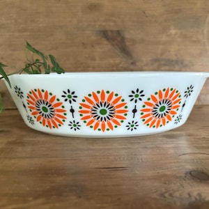 May include: White oval casserole dish with orange and green floral pattern. The dish has a handle on each side. The floral design features orange petals with green centers and black accents. A sprig of greenery is in the background.