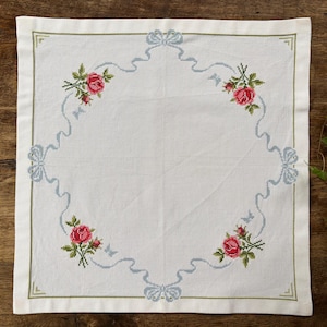 Peut inclure: Un carré de tissu blanc orné d'un motif au point de croix représentant quatre bouquets de roses rouges aux coins, reliés par un ruban bleu clair avec des nœuds. Une fine bordure verte encadre l'ensemble du carré de tissu.