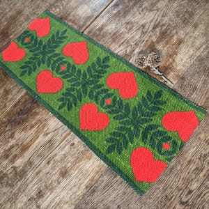 Puede incluir: Un camino de mesa rectangular de tela verde con un patrón repetido de corazones rojos y hojas verde oscuro. El camino de mesa está colocado sobre una superficie de madera. El diseño del camino de mesa es un motivo encantador inspirado en la naturaleza.