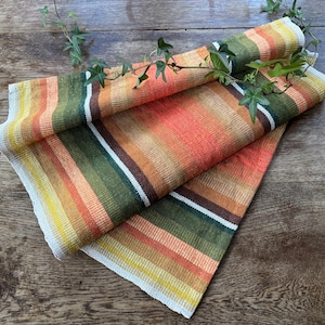 Könnte beinhalten: Gefaltetes Textil mit einem Streifenmuster in Orange-, Grün-, Gelb- und Weißtönen. Der Stoff ist mit einem Zweig aus grünen Blättern verziert. Das Textil liegt auf einer Holzoberfläche.