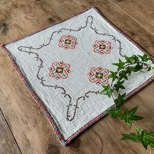 May include: A white square table runner with a brown, orange, and blue floral pattern. The runner has a white background with a brown border and a floral design in the center. The runner is on a wooden surface.