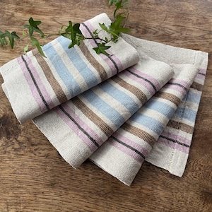 May include: A stack of linen napkins with a striped pattern in light blue, brown, pink, and black. The napkins are folded and arranged on a wooden surface, with a sprig of greenery in the background.