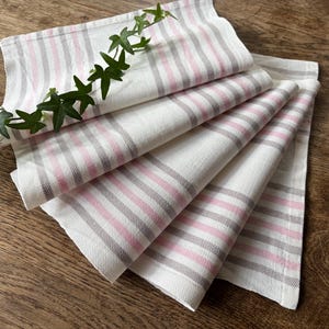 Puede incluir: Servilletas de tela blancas dobladas con rayas rosas y grises. Las servilletas están dispuestas en forma de abanico, con una ramita de hojas verdes encima. Las servilletas son de tela suave y texturizada.