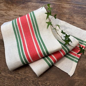 Op de afbeelding: Gevouwen linnen servetten met rode en groene strepen op een crèmekleurige achtergrond. De servetten zijn versierd met een takje groene bladeren. De servetten liggen op een houten oppervlak.